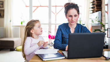 Woman working on laptop while her small daughter stands next to her