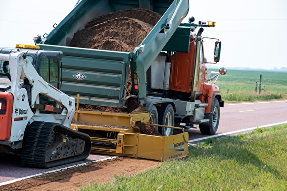 Truck dumping gravel onto a road shoulder using the road widener attachment