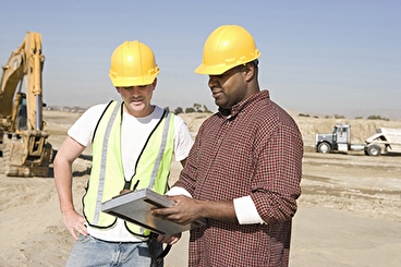 Two male construction workers, one Black and one white, wearing hard hats and looking at plans