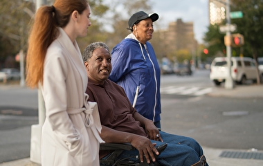 Three people on a sidewalk near an intersection. The one in the middle is using a wheelchair.