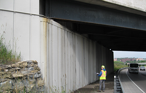 MnDOT intern Tristan Karls inspecting a bridge wall while traffic drives by