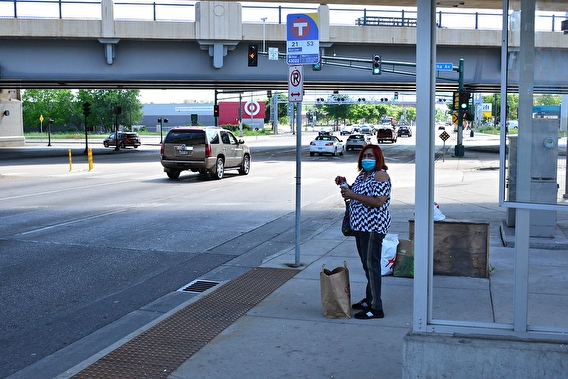 Woman wearing a mask standing at a bus stop with a grocery bag at her feet