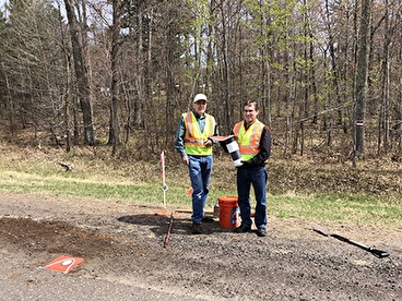 Two white men wearing safety vests and holding equipment, outside on gravel road in woods